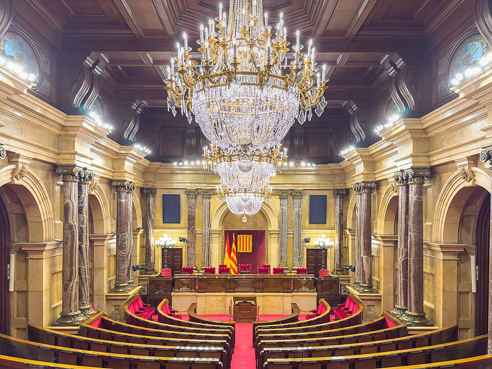 Hemicicle del Parlament de Catalunya amb una gran aranya de llum al centre i els escons vermells. Fotografia de Kent Wang (CC BY-SA 4.0, Wikimedia Commons).
